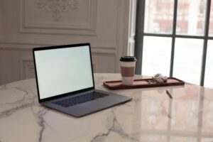 A minimalist interior setup featuring a laptop, coffee cup, and pen on a marble table by a window.